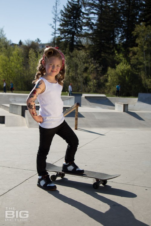 children's portraits, little skater girl with retro hair and fake tattoo sleeve standing on a skateboard in a skate park on a sunny day, children's photography, skater girl