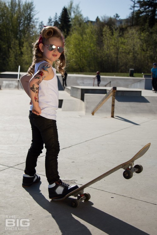 children's portraits, little skater girl with sunglasses and retro hair and fake tattoo sleeve standing on a skateboard with her hands on her hips in a skate park on a sunny day, children's photography, skater girl