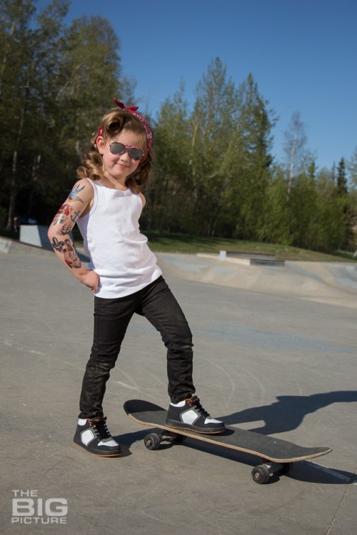 children's portraits, smiling little skater girl wearing sunglasses with retro hair and fake tattoo sleeve standing on a skateboard in a skate park on a sunny day, children's photography, skater girl