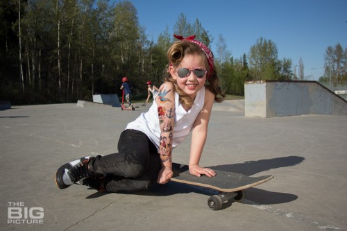 children's portraits, smiling little skater girl wearing sunglasses with retro hair and fake tattoo sleeve sitting on a skateboard in a skate park on a sunny day, children's photography, skater girl