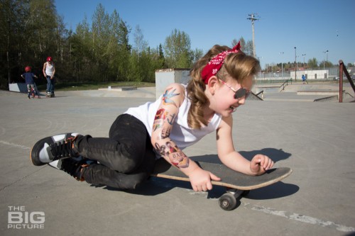 children's portraits, smiling little skater girl wearing sunglasses with retro hair and fake tattoo sleeve laying on a skateboard in a skate park on a sunny day, children's photography, skater girl