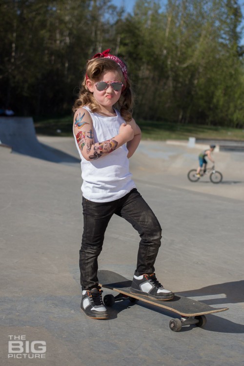 children's portraits, smiling little skater girl wearing sunglasses with retro hair and fake tattoo sleeve standing on a skateboard flexing her muscles in a skate park on a sunny day, children's photography, skater girl