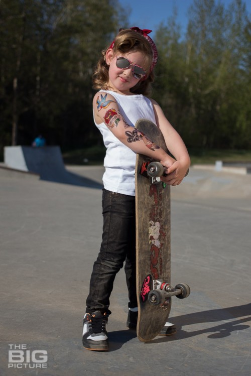 children's portraits, smiling little skater girl wearing sunglasses with retro hair and fake tattoo sleeve holding a skateboard in a skate park on a sunny day, children's photography, skater girl